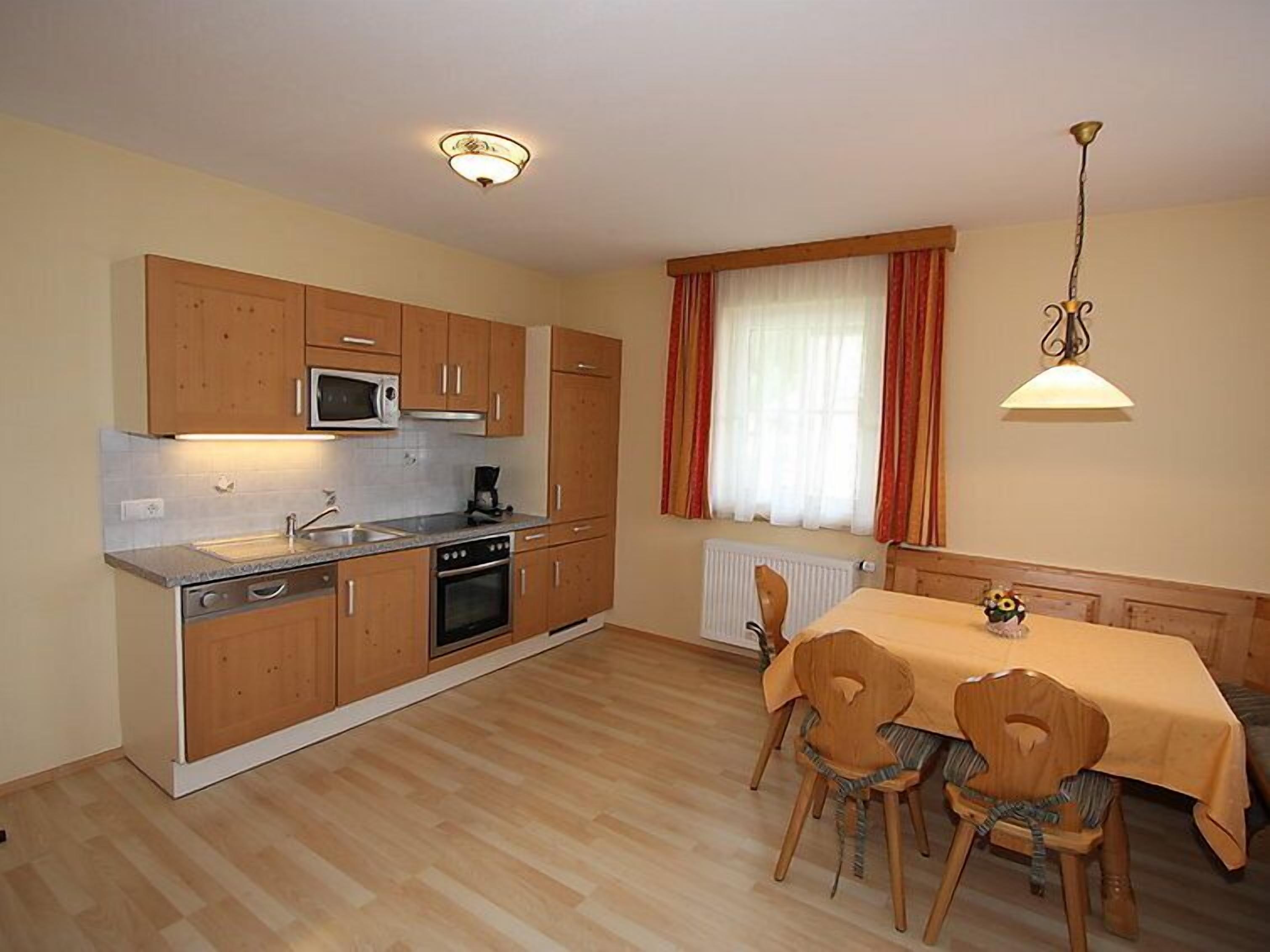 Una cocina compacta con armarios de madera, microondas, lavamanos y estufa. Hay una mesa de comedor con sillas y una ventana con cortinas.