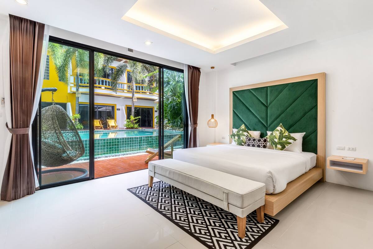 A modern bedroom with a large bed, a bench, a bedside table, and a view of a pool and outdoor area.
