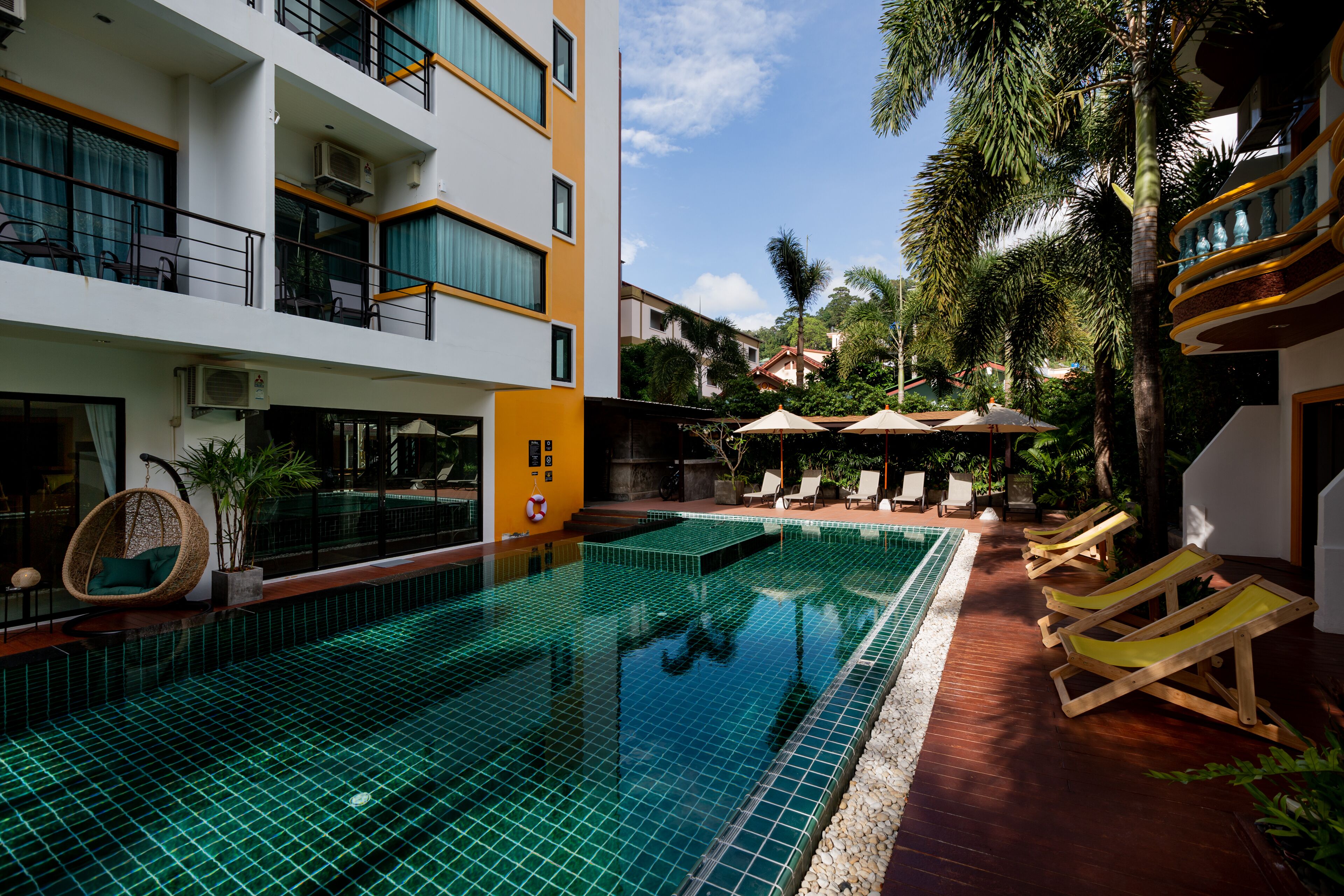 A swimming pool with lounge chairs and umbrellas, surrounded by buildings and palm trees.
