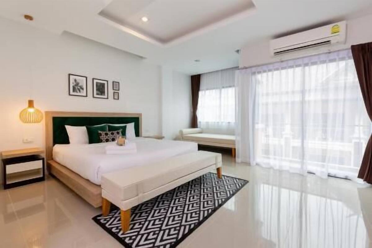 A modern hotel room with a large bed, bedside tables, a bench, a patterned rug, and a large window with curtains.