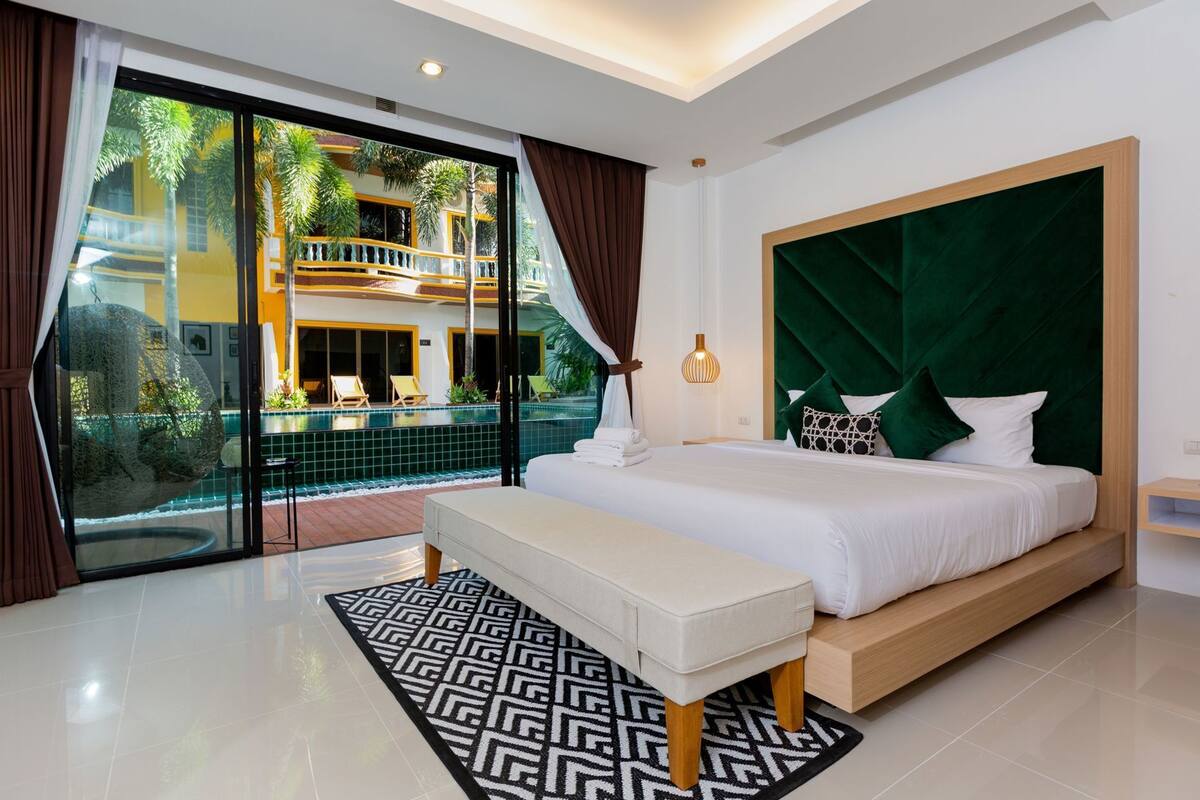 A bedroom with a large bed, a bench, a view of a pool and building, and a green headboard.