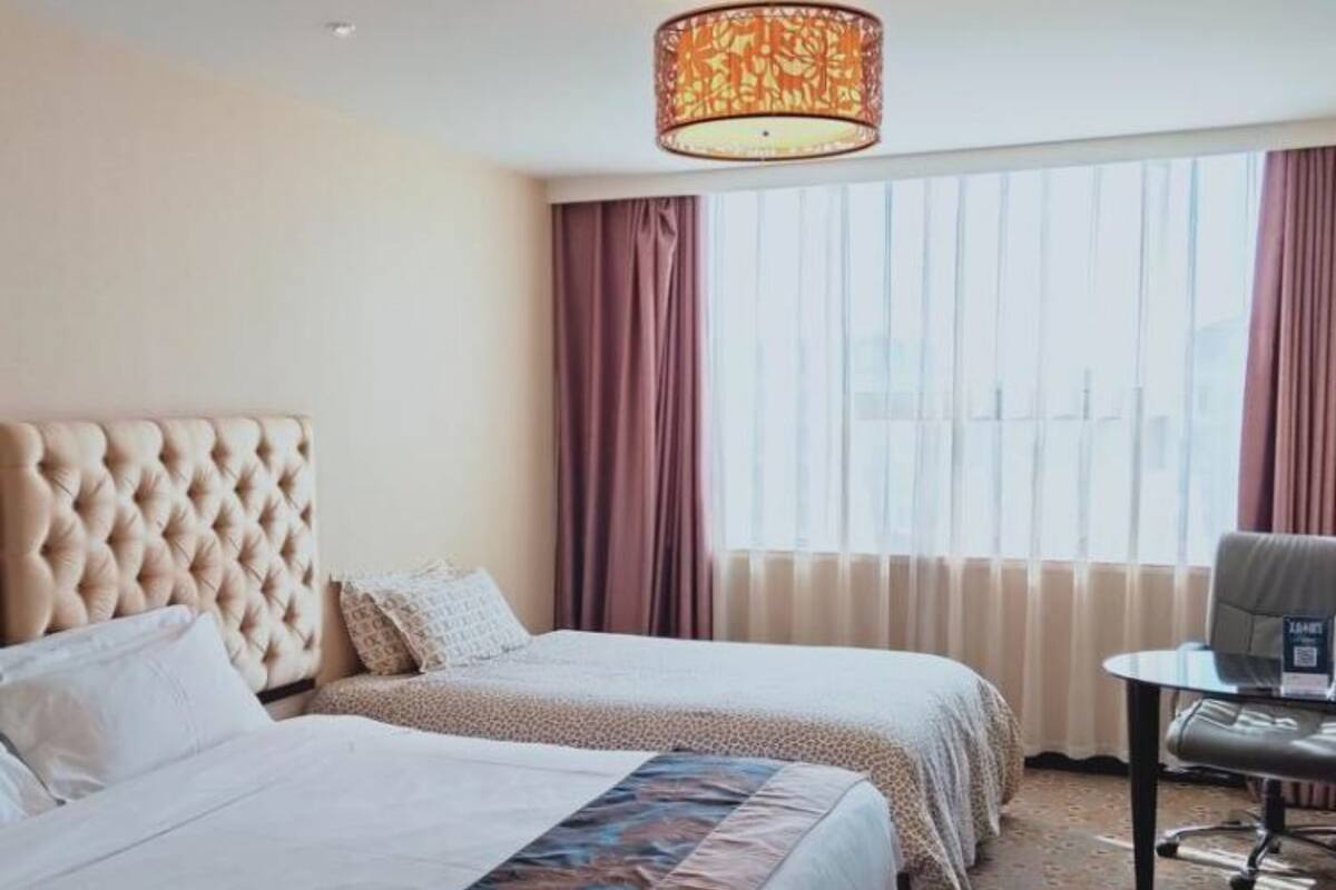 Une chambre d’hôtel avec deux lits, une tête de lit capitonnée, une couvre-lit à motifs, un bureau avec une chaise et une grande fenêtre avec des rideaux.