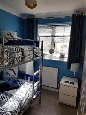 A bunk bed room with a window, a radiator, a bedside table with a lamp, and a wall with a picture frame.