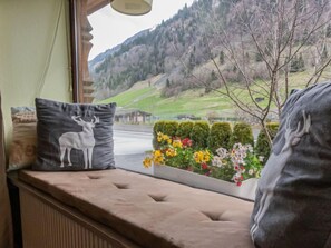 Asiento junto a la ventana con un cojín que tiene un diseño de ciervo, una maceta con flores de colores y vistas a un paisaje montañoso.