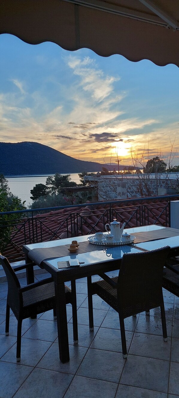 A terrace with a table set for two, overlooking a body of water and a distant mountain range during sunset.