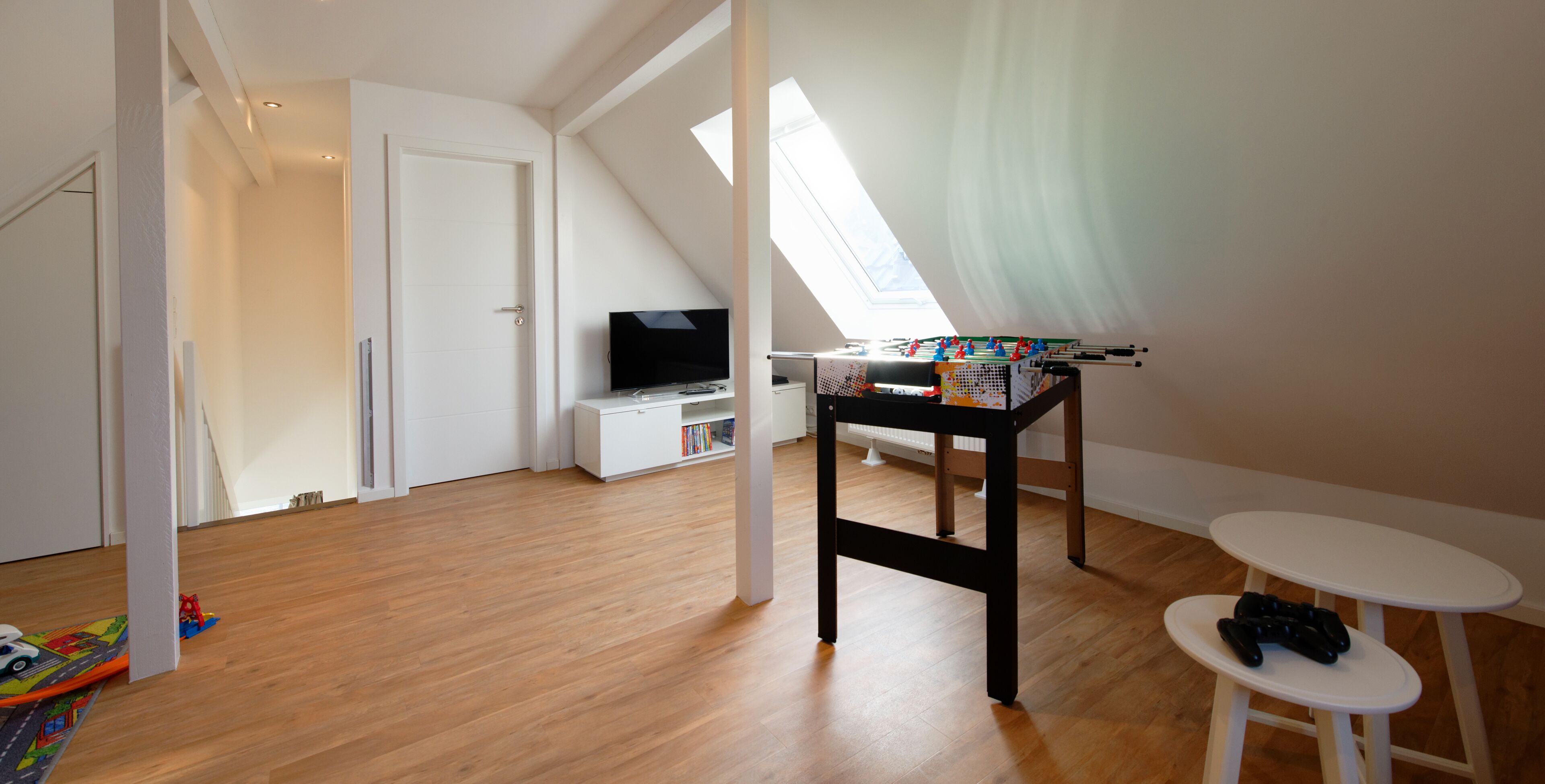 Una habitación con suelo de madera, una puerta blanca, un televisor sobre un mueble blanco, una mesa de fútbol de mesa y una pequeña mesa blanca.