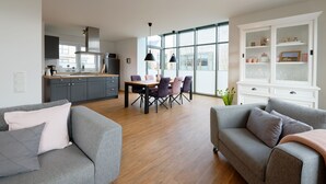 Un salon moderne avec des canapés gris, une table à manger et un coin cuisine doté de meubles gris et d’une fenêtre.