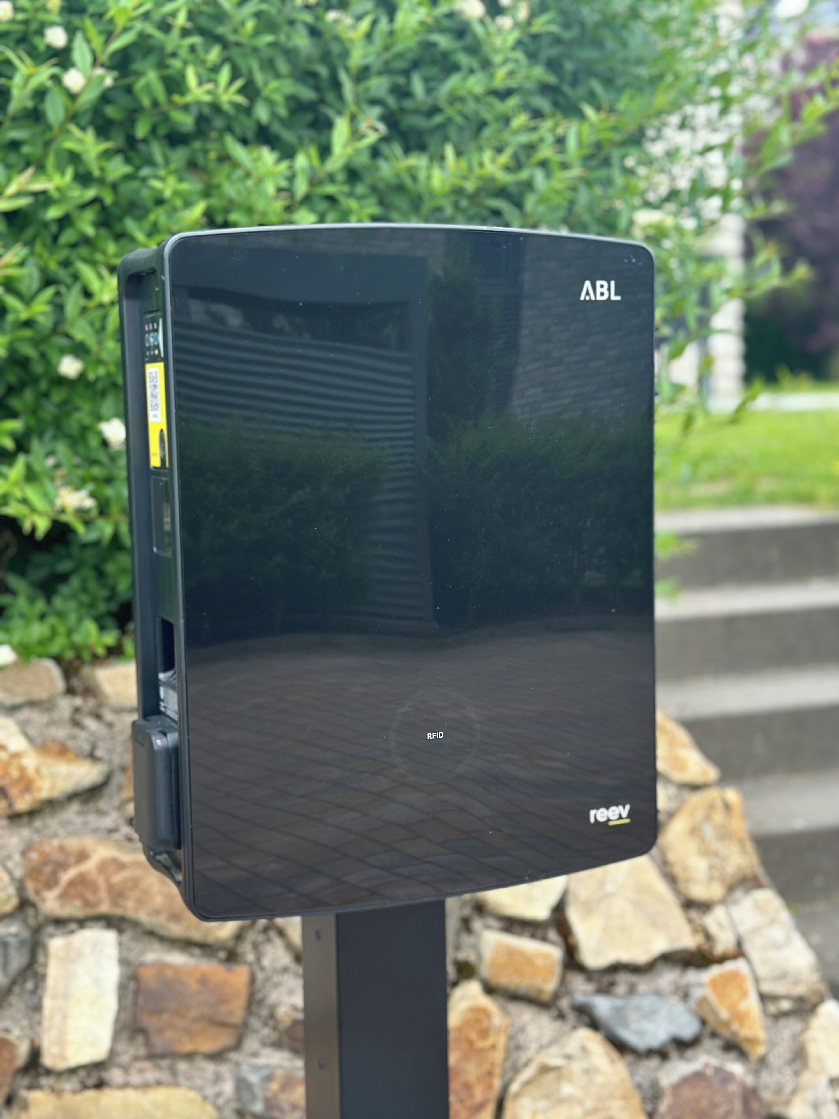 Ein schwarzes Outdoor-WLAN-Gerät mit 'reev'-Branding, an einem Metallpfosten befestigt, umgeben von einer Steinmauer und Pflanzen.