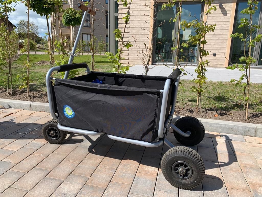 A black wheeled cart with a logo, parked on a paved surface with young trees and a building in the background.