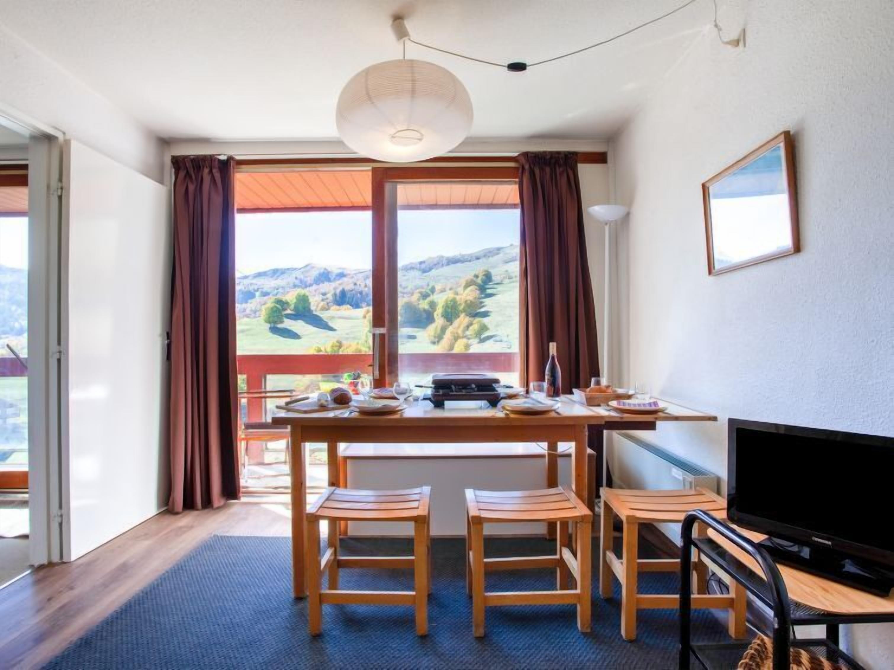 A room with a wooden dining table, four chairs, a television, and a view of a mountainous landscape through large windows.