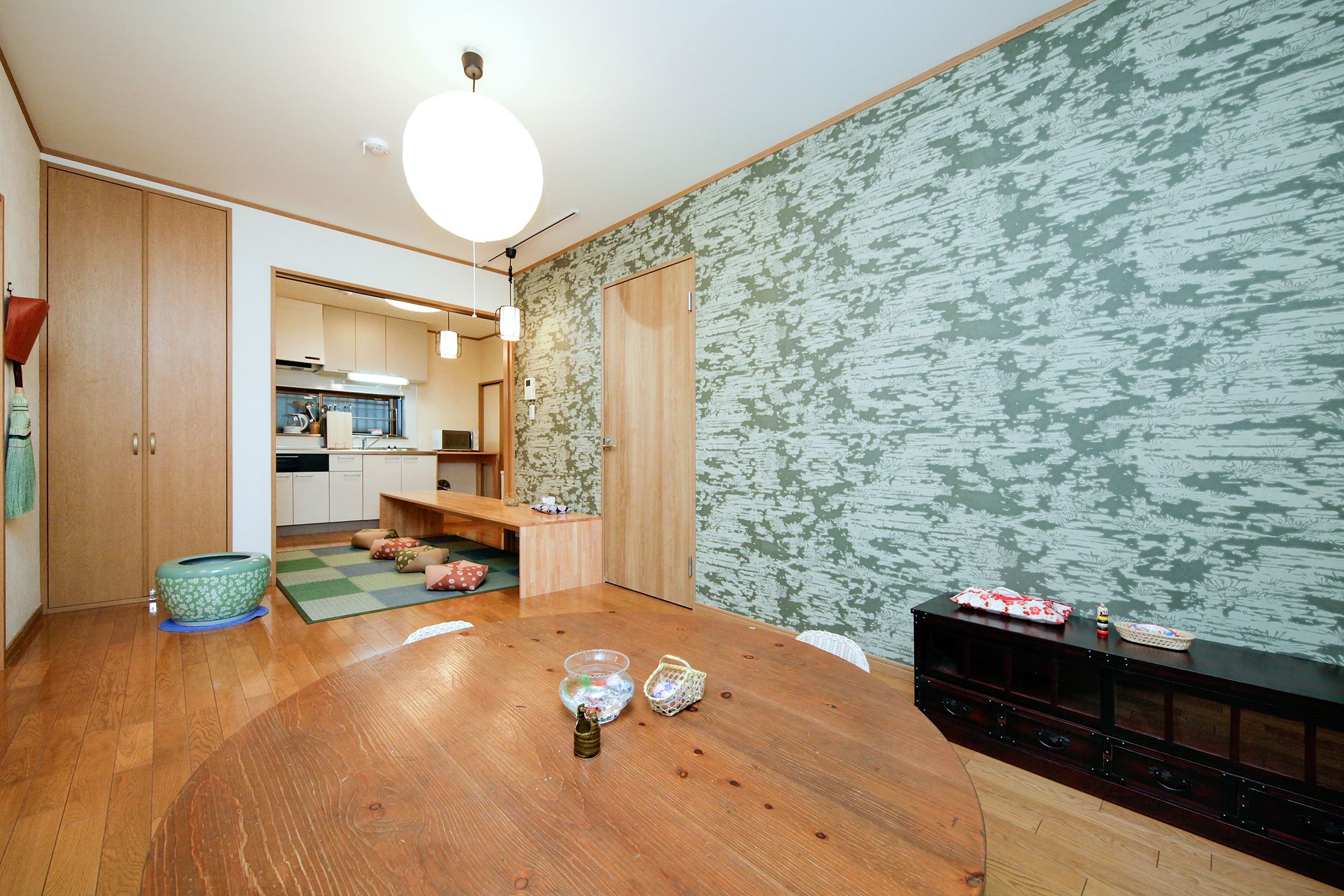 A dining area with a wooden table, a kitchen in the background, and a patterned wall.