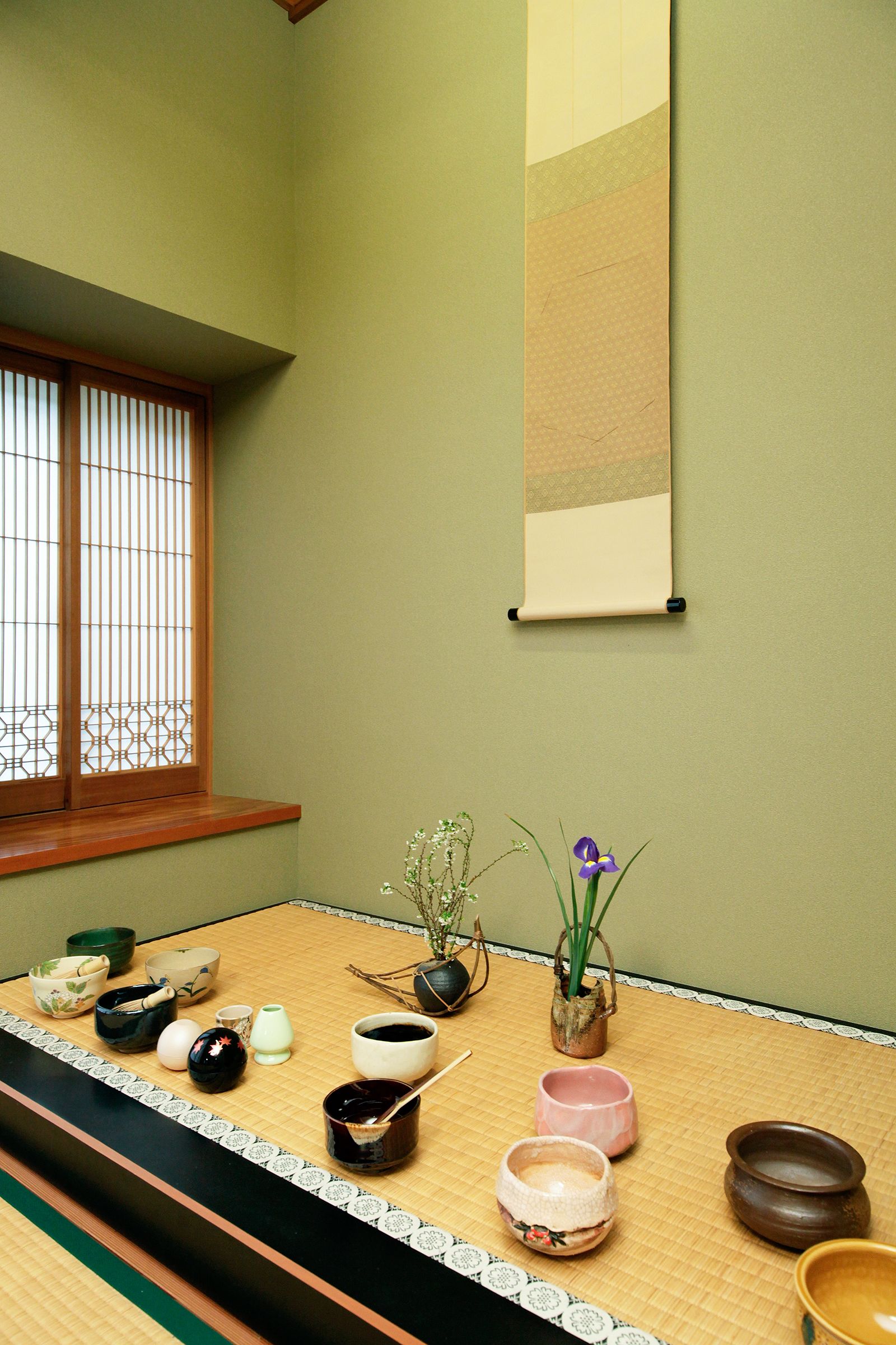A tatami mat room with a low table, various bowls, a hanging scroll, and a window with wooden frames.