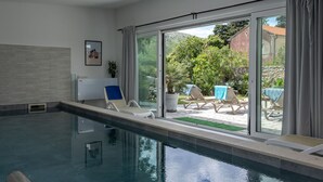 Ein moderner, überdachter Schwimmbereich mit Glaswänden, einer Deckenleuchte und Liegestühlen mit Handtüchern.