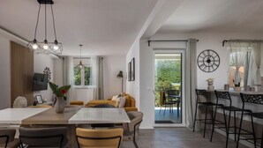 Ein modernes Wohnzimmer mit Essbereich, einer Couch, einem Fernseher und einem Balkon mit Blick ins Grüne.