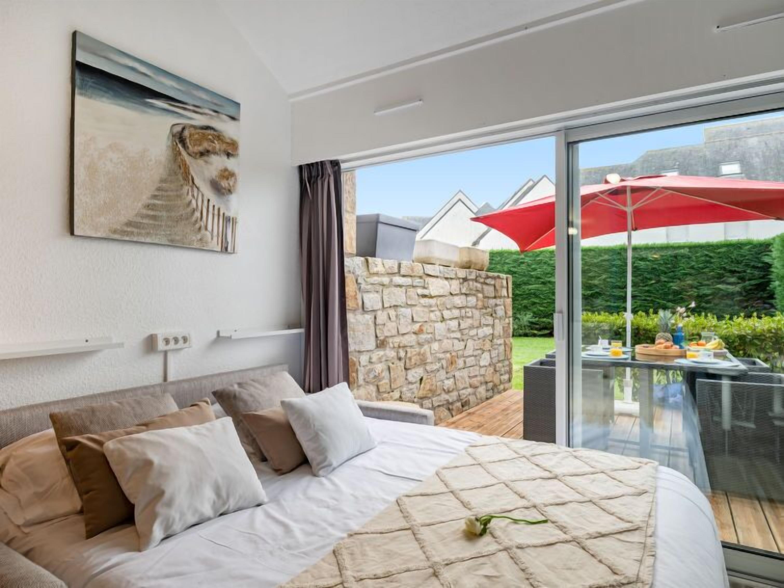 A bedroom with a large bed, a stone wall, and a glass balcony with a red umbrella.
