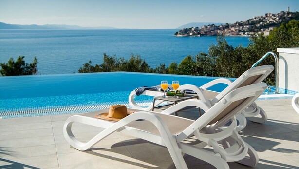 Ein Poolbereich mit Liegestühlen, einem Tisch mit Getränken und einem Hut, mit Blick auf das Meer und eine in der Ferne liegende Stadt.