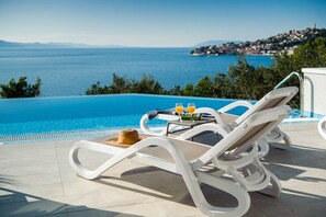 Un espace aménagé au bord de la piscine, avec des chaises longues, une table où sont posées des boissons et un chapeau, et une vue sur la mer et une ville située au loin.