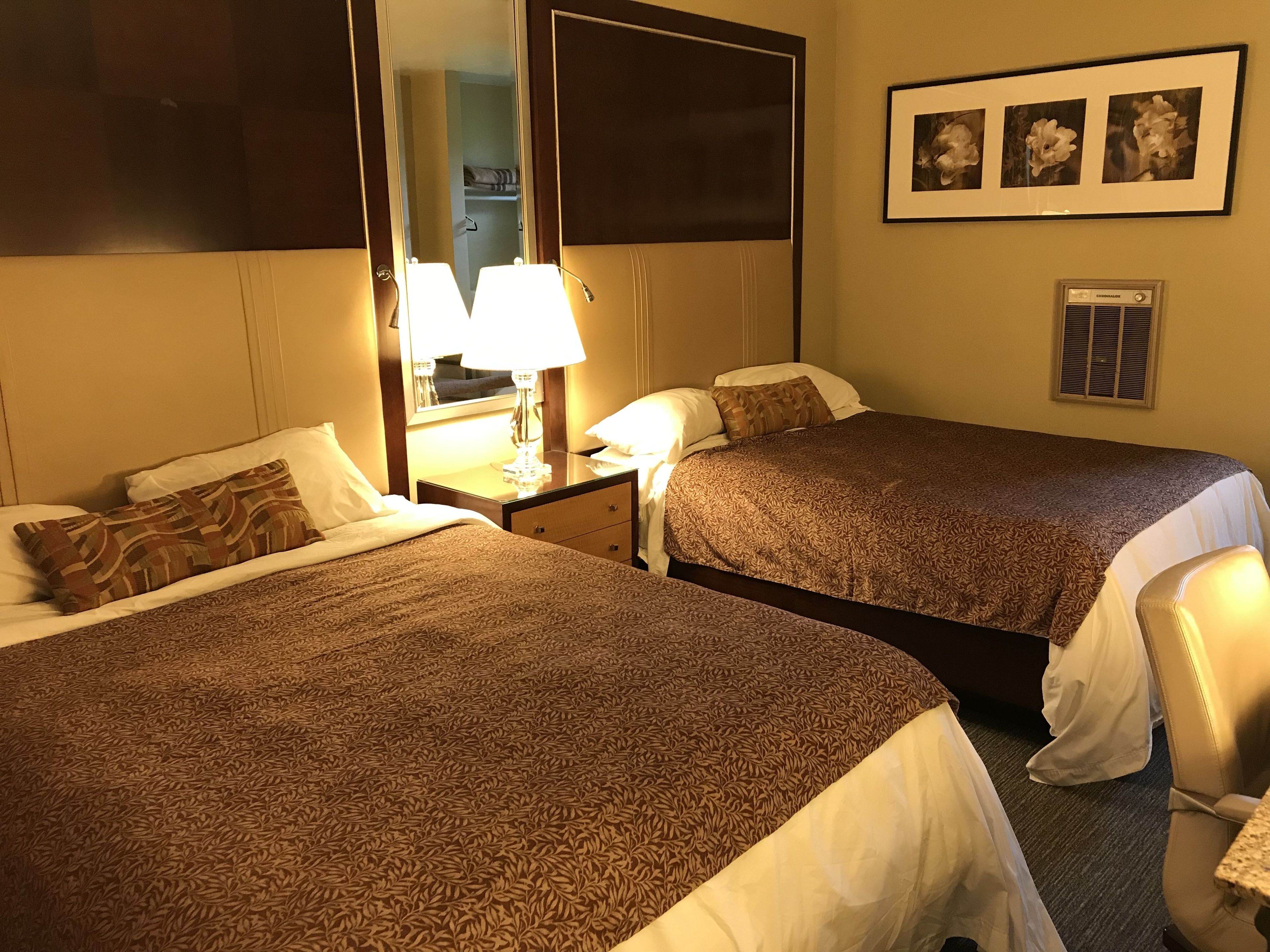 A hotel room with two beds, a nightstand with a lamp, a mirror, and a framed picture on the wall.
