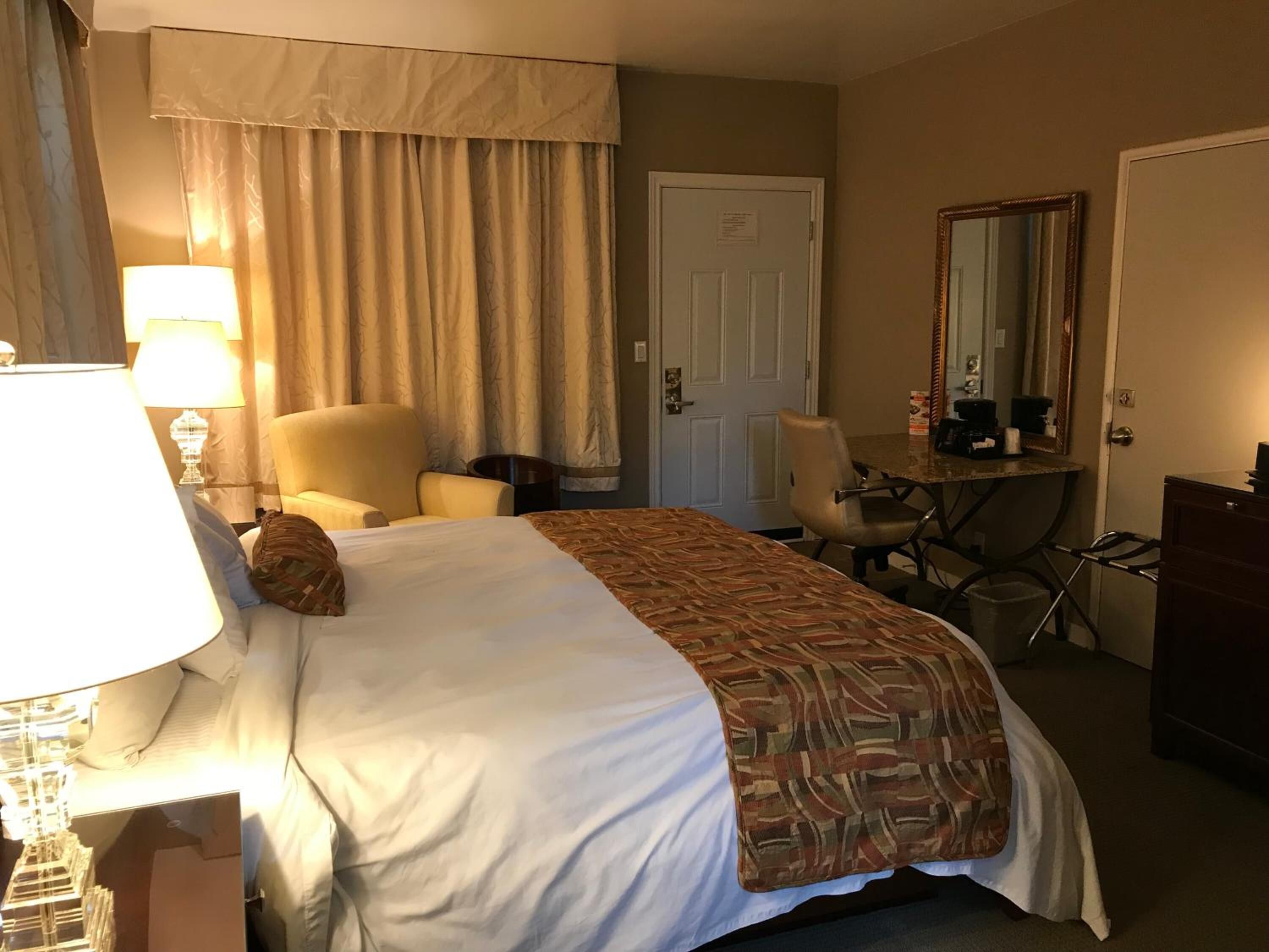Une chambre d’hôtel avec un lit, un bureau, une chaise, une lampe, un miroir et une porte.