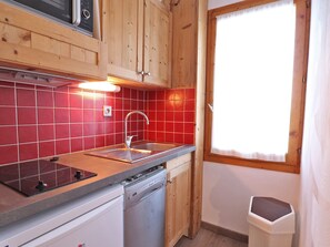 Une cuisine compacte avec des meubles en bois, un crédence en carreaux rouges, un évier en acier inoxydable et une fenêtre agrémentée d’un rideau blanc.