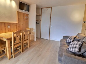 A room with a wooden dining table, chairs, a sofa with plaid pillows, a television mounted on the wall, and a wardrobe with a door.