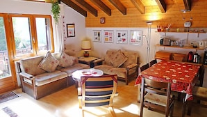 Una sala de estar acogedora con vigas de madera, un conjunto de sofá, una silla a rayas, una mesa de comedor con mantel rojo y una zona de cocina con estantes y electrodomésticos.