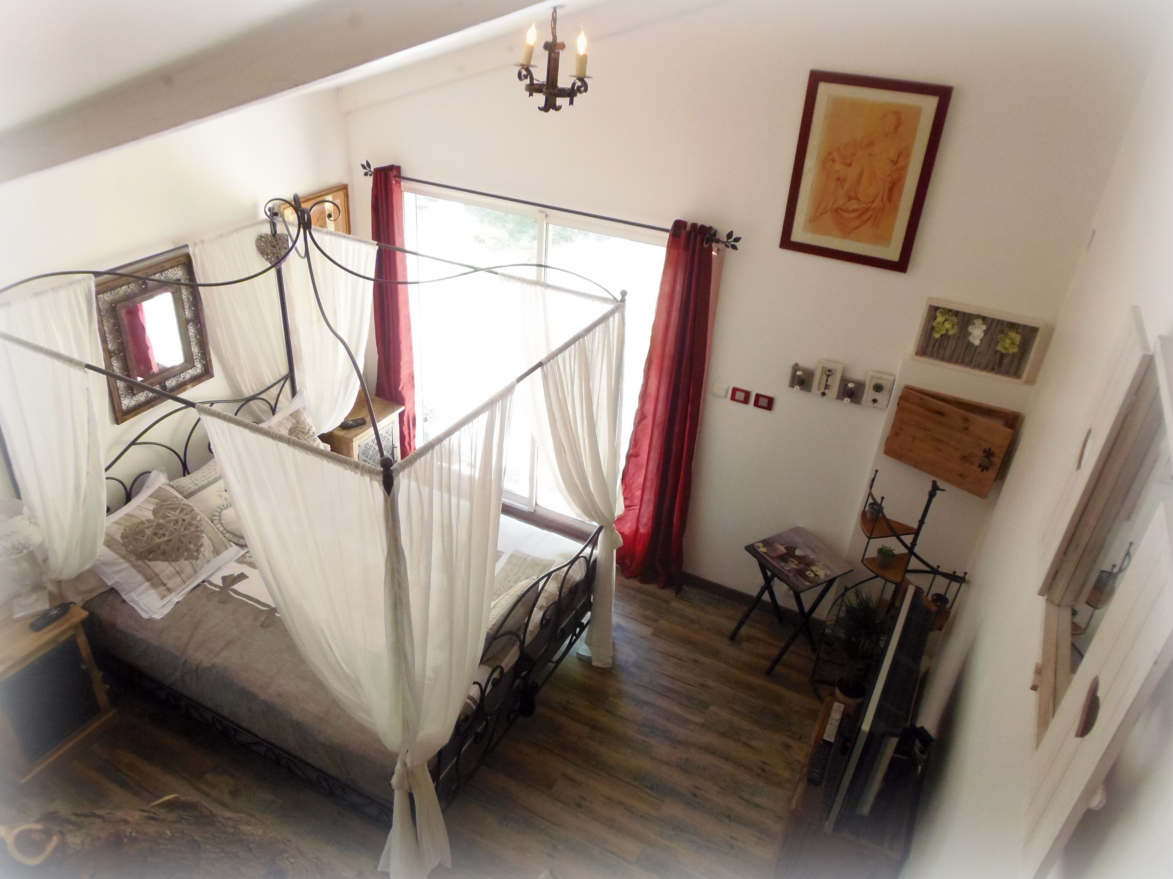 A bedroom with a four-poster bed, a chandelier, a mirror, and a window with red curtains.