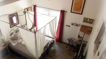 A bedroom with a four-poster bed, a chandelier, a mirror, and a window with red curtains.