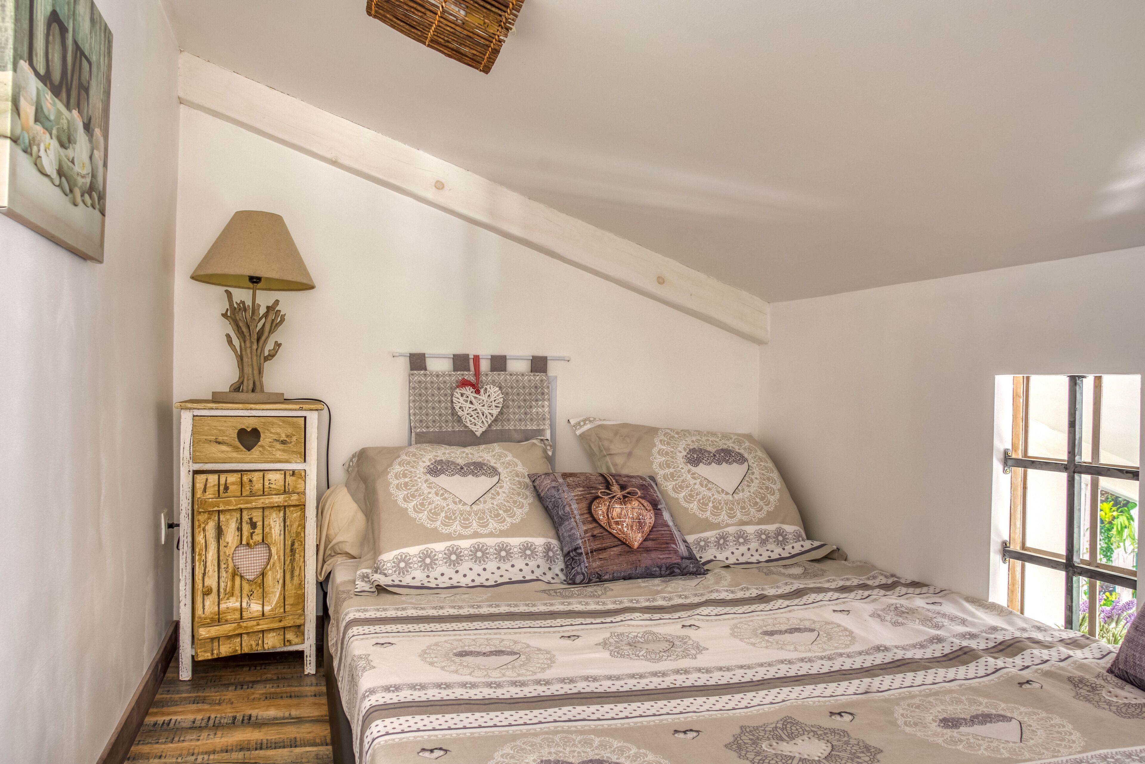 A bedroom with a bed, a wooden chest of drawers, a lamp, and a window with a view of greenery.