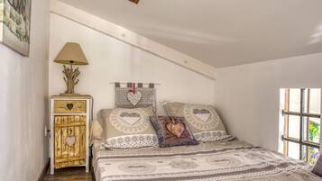 A bedroom with a bed, a wooden chest of drawers, a lamp, and a window with a view of greenery.