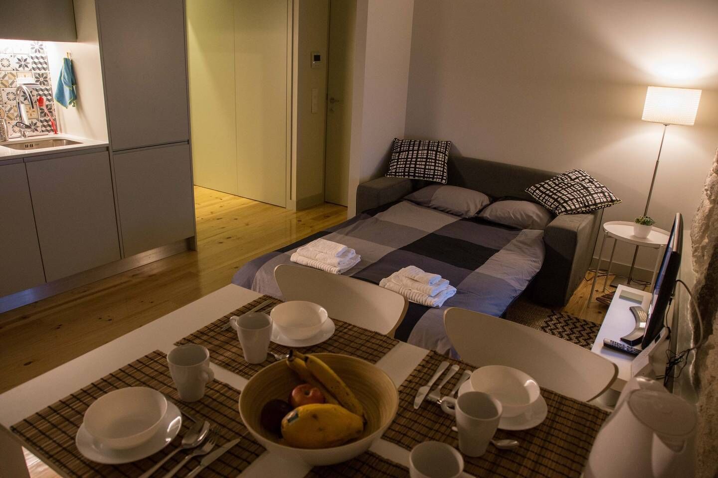 Habitación de hotel compacta con cama, pequeño espacio para sentarse, kitchenette y mesa de comedor con un tazón de fruta.