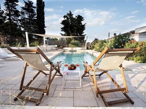 A poolside area with two wooden lounge chairs, a small table with fruit and drinks, and a view of the pool and surrounding greenery.