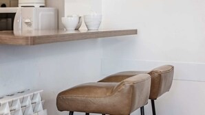 Une cuisine avec une étagère en bois sur laquelle sont posés un appareil à café, deux bols blancs et un ensemble de tasses blanches. On y trouve également deux chaises en cuir marron avec des pieds noirs.