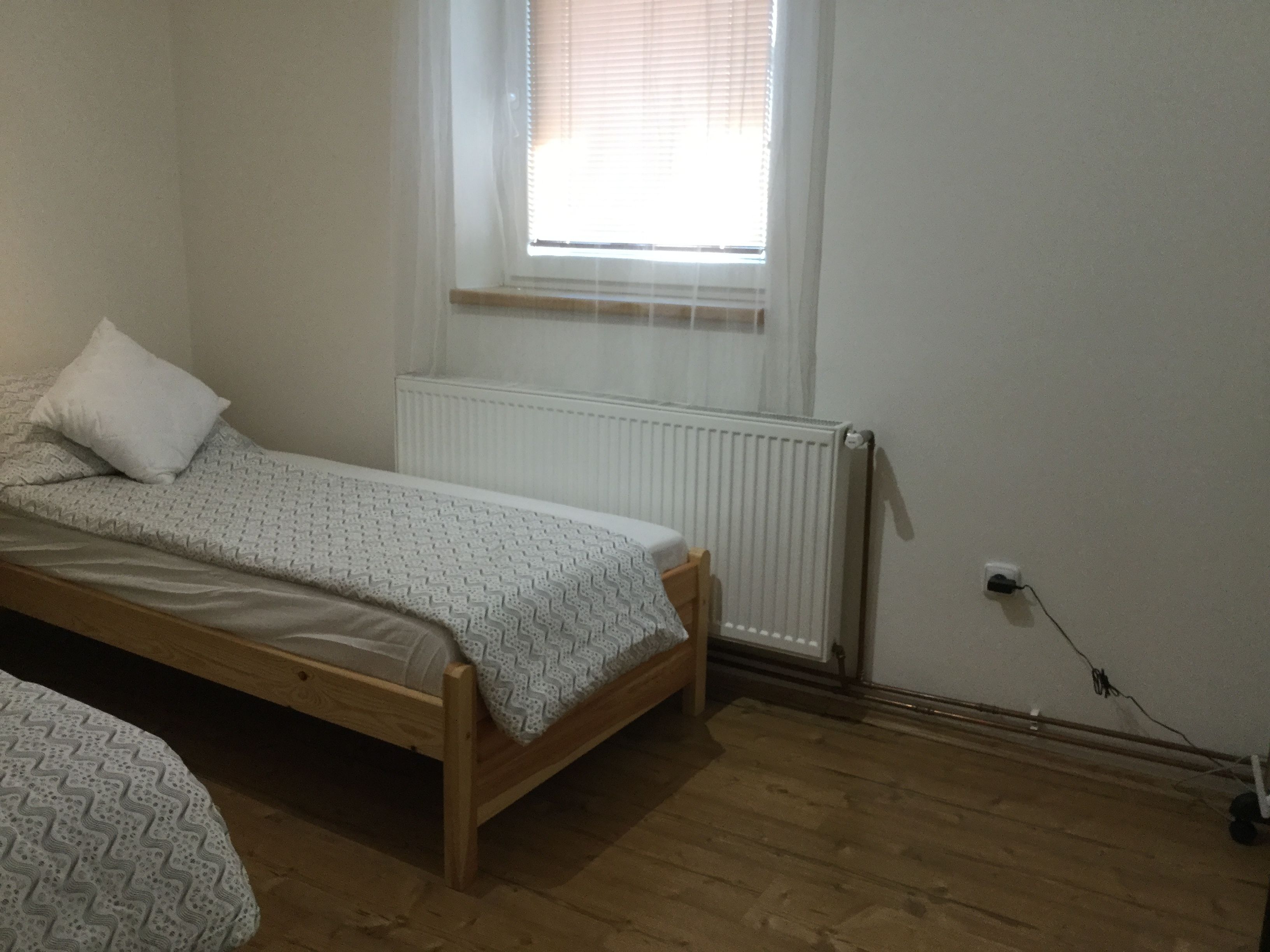 A small, single-bed room with a wooden floor, a radiator, and a window with blinds.