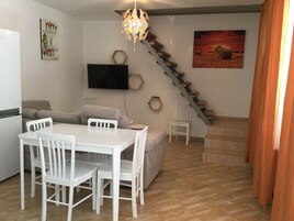 Una sala de estar moderna con una mesa de comedor, sillas blancas, un sofá, un televisor montado en la pared, una escalera y un cuadro.