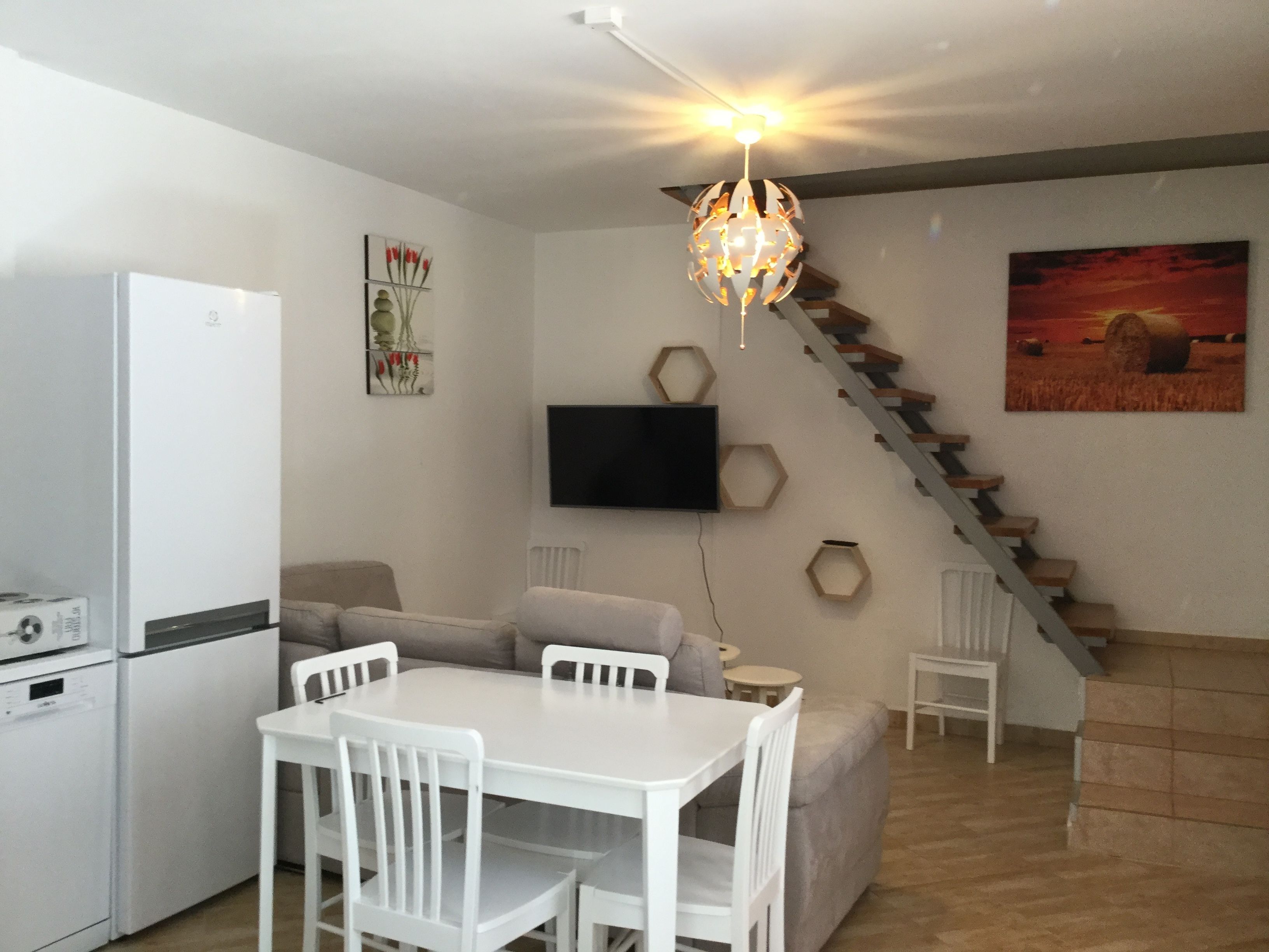 Un espacio habitable compacto con cocina, una mesa de comedor con sillas, un sofá, un televisor montado en la pared y una escalera.