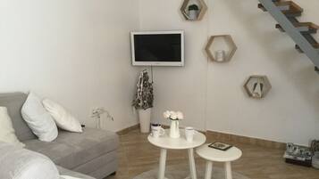 Un salon avec un canapé gris, une petite table blanche, un téléviseur fixé au mur et un escalier en bois.
