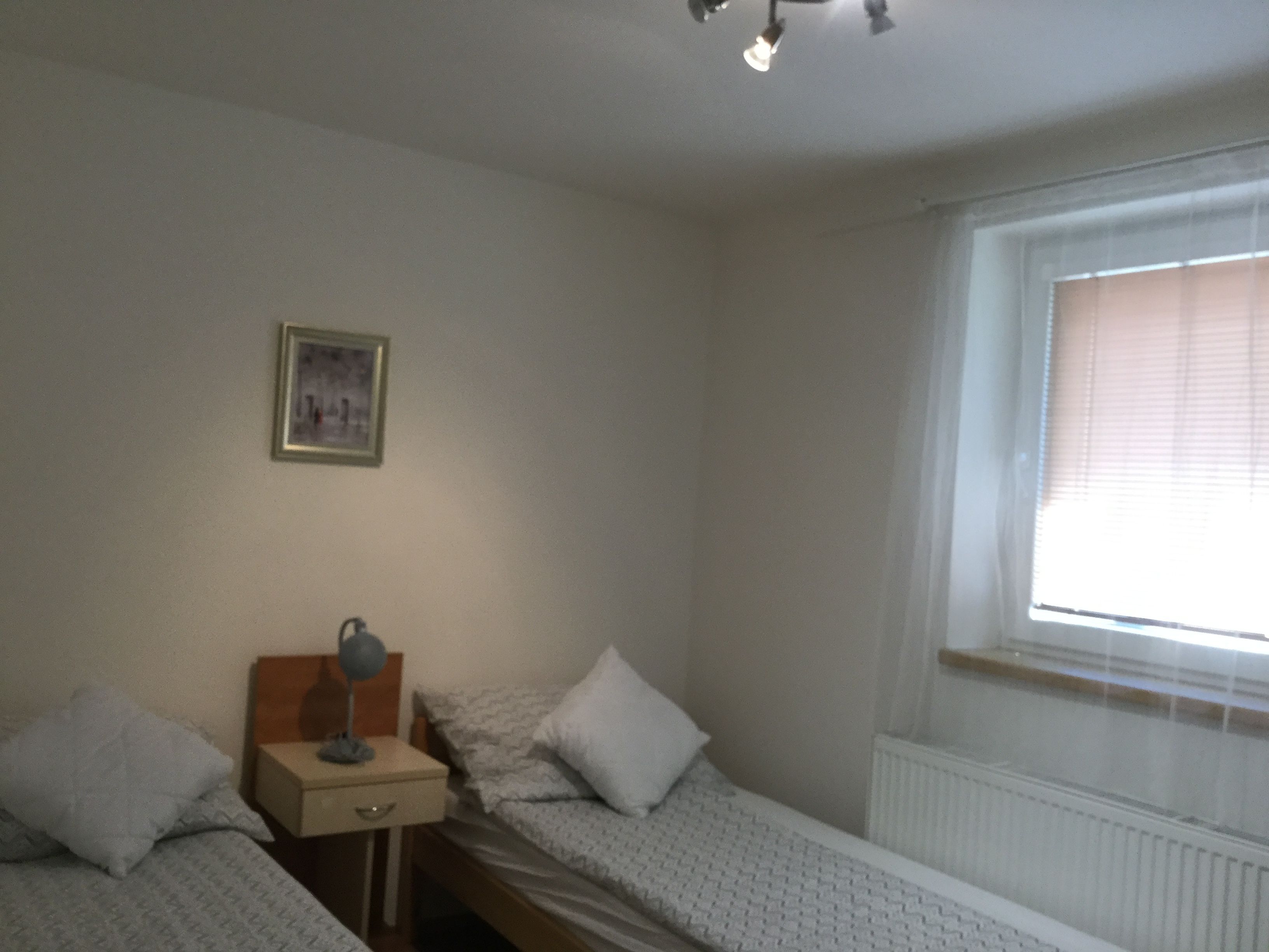 Ein schlichter, kleiner Raum mit zwei Einzelbetten, einem Nachttisch mit Lampe, einem gerahmten Bild an der Wand und einem Fenster mit Jalousien.