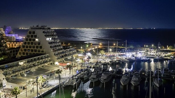 Een haven 's nachts met aangemeerde boten, een groot gebouw met meerdere balkons en een rustige zee onder een heldere maan.