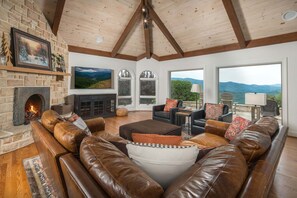 A cozy living room with a fireplace, leather sofas, and large windows offering a view of the outdoors.
