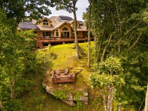 A two-story house with a deck and solar panels, surrounded by trees and a grassy yard.