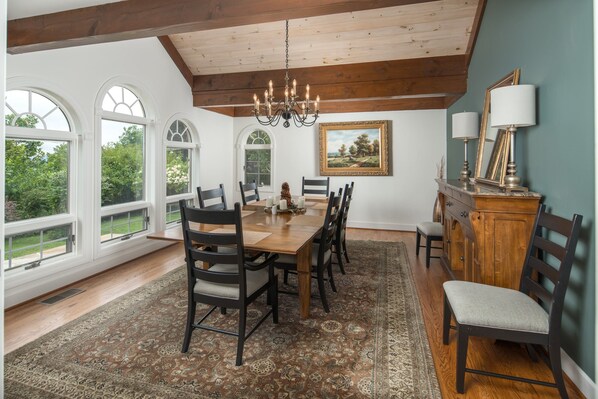 Comedor con mesa de madera, sillas, una lámpara de araña y vistas a la vegetación a través de grandes ventanales.