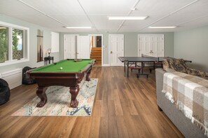 Una habitación con mesa de billar, mesa de ping pong, un sofá y una escalera que sube a un segundo piso.
