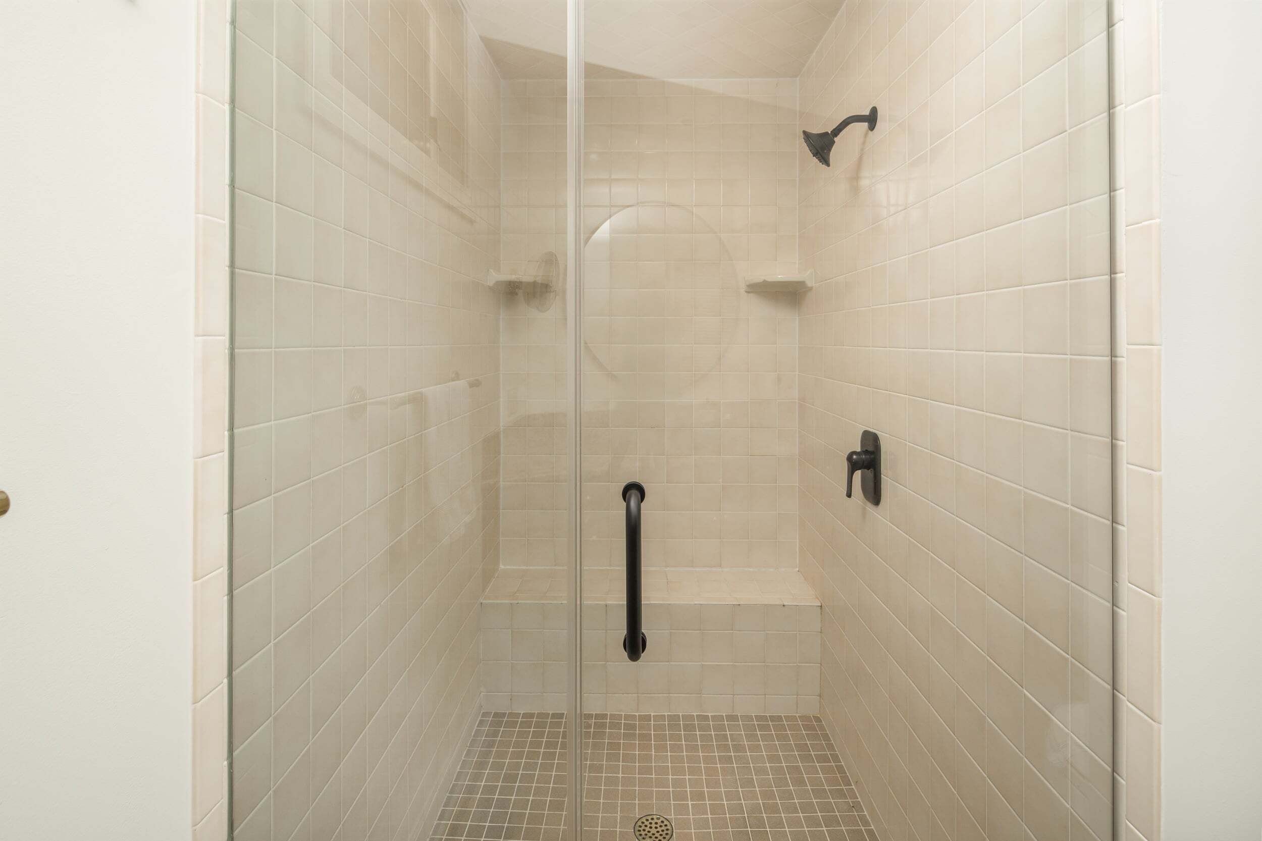 A glass-enclosed shower stall with beige tiles, a handheld showerhead, and a wall-mounted faucet.