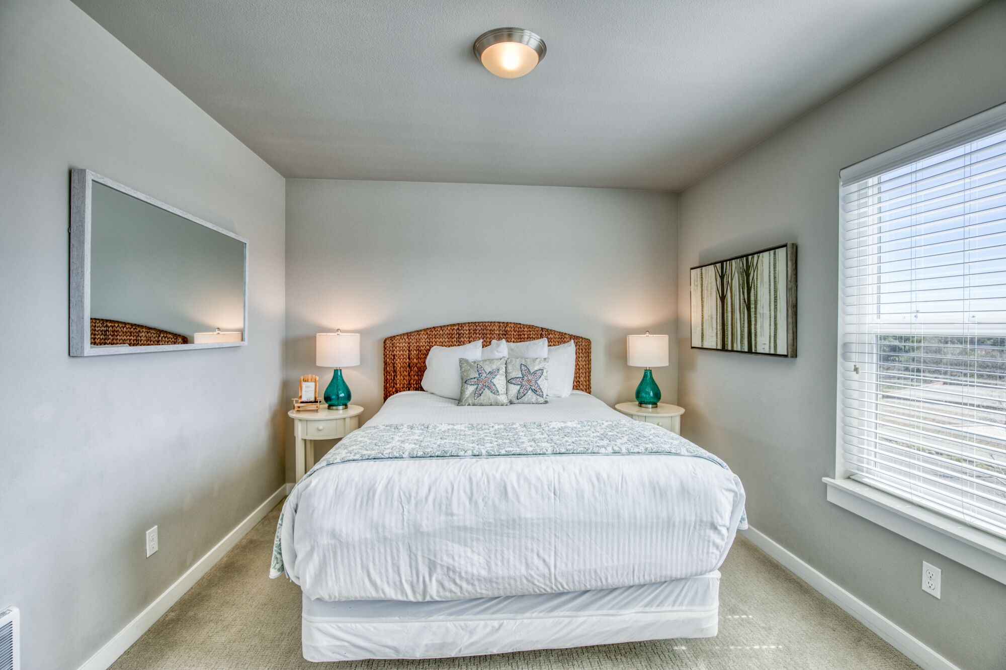 Une chambre avec un grand lit, deux tables de chevet avec des lampes, un luminaire au plafond, une grande fenêtre avec des stores et un miroir fixé au mur.
