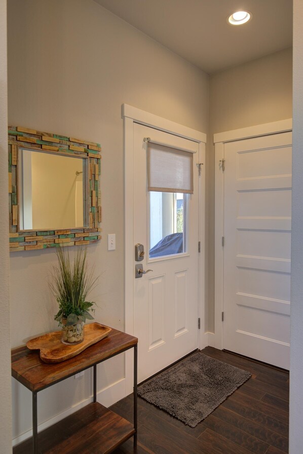 A hallway with a white door, a window with a blind, a mirror with a wooden frame, and a small table with a plant.
