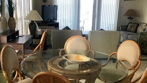 Un comedor con mesa de vidrio rodeada de sillas de mimbre, una araña y un televisor al fondo.