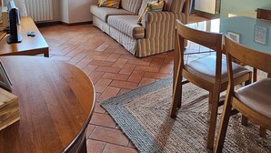 Ein gemütliches Wohnzimmer mit Holzboden, einem gestreiften Sofa, einem Esstisch und Blick ins Freie durch große Fenster.