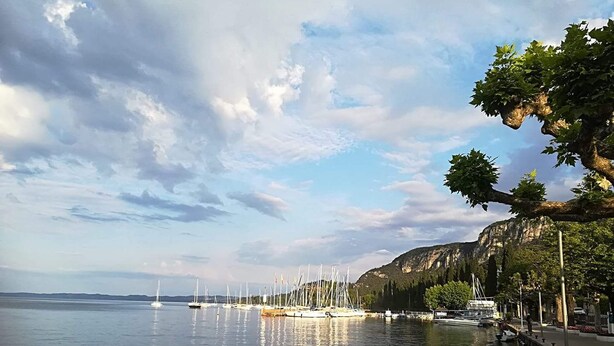 Ruhiges Wasserufer mit vor Anker liegenden Booten, einer Promenade und einer Bergkulisse im Hintergrund.