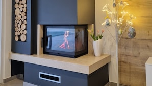 A modern living room with a built-in fireplace, a wall-mounted TV, and decorative elements like a wooden wall and a plant with lights. - Villa**** SANIVARO with heated pool and SPA (Sanvincenti)